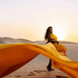 May include: A woman in a flowing yellow dress stands in a desert landscape at sunset. The dress fabric billows in the wind, creating a dynamic composition against the backdrop of sand dunes and a warm, golden sky.