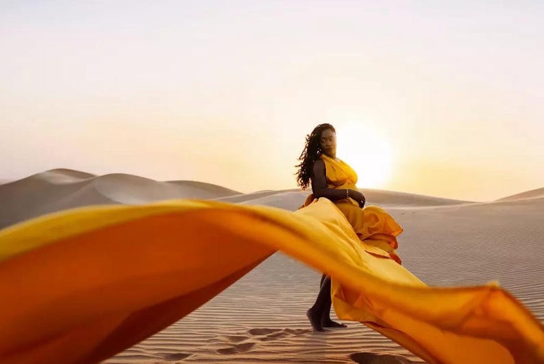 May include: A woman in a flowing yellow dress stands in a desert landscape at sunset. The dress fabric billows in the wind, creating a dynamic composition against the backdrop of sand dunes and a warm, golden sky.