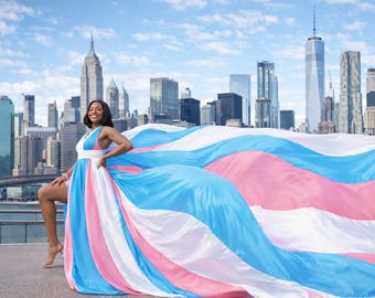 May include: A woman in a flowing dress with blue, pink, and white panels stands in front of a city skyline. The dress's colors mirror the transgender pride flag, creating a vibrant and symbolic image.
