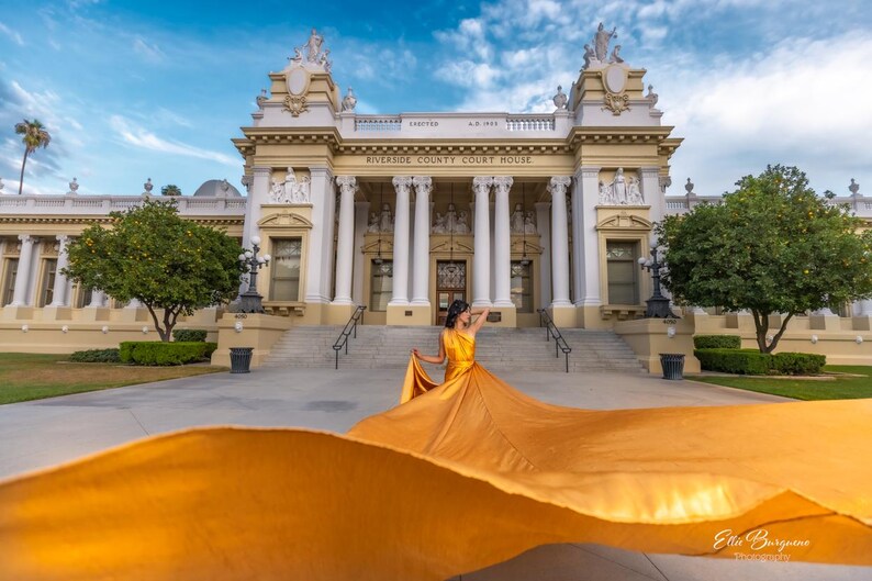 May include: A woman in a flowing, golden-yellow dress poses in front of the Riverside County Courthouse. The building features white columns and a cream-colored facade under a blue sky. The dress fabric billows dramatically.