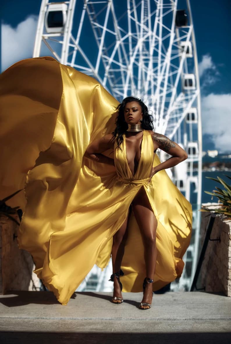 May include: A woman in a flowing, golden yellow dress stands in front of a Ferris wheel. The dress has a plunging neckline and a high slit, revealing the legs. She wears a gold choker and high-heeled sandals. The fabric billows in the wind.