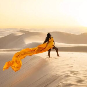 May include: A woman in a yellow dress stands on a sand dune, holding a long, flowing yellow fabric. The desert landscape features rolling sand dunes under a warm, golden sky. The image captures a moment of grace and beauty.