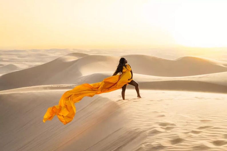 May include: A woman in a yellow dress stands on a sand dune, holding a long, flowing yellow fabric. The desert landscape features rolling sand dunes under a warm, golden sky. The image captures a moment of grace and beauty.