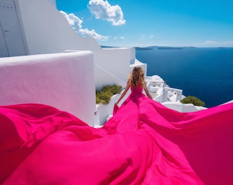 May include: A woman in a flowing, hot pink dress walks along a white building overlooking the ocean. The dress billows out behind her, creating a dramatic effect against the blue sky and sea. The architecture is classic, with white walls and steps.