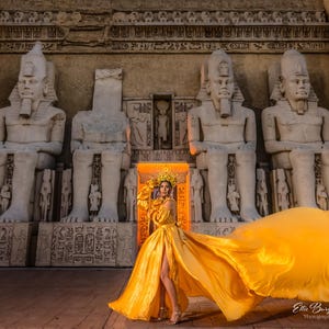 May include: A woman in a flowing gold dress and headdress stands before ancient Egyptian statues. The dress has a high slit and a long train. The background features large stone statues and hieroglyphs.
