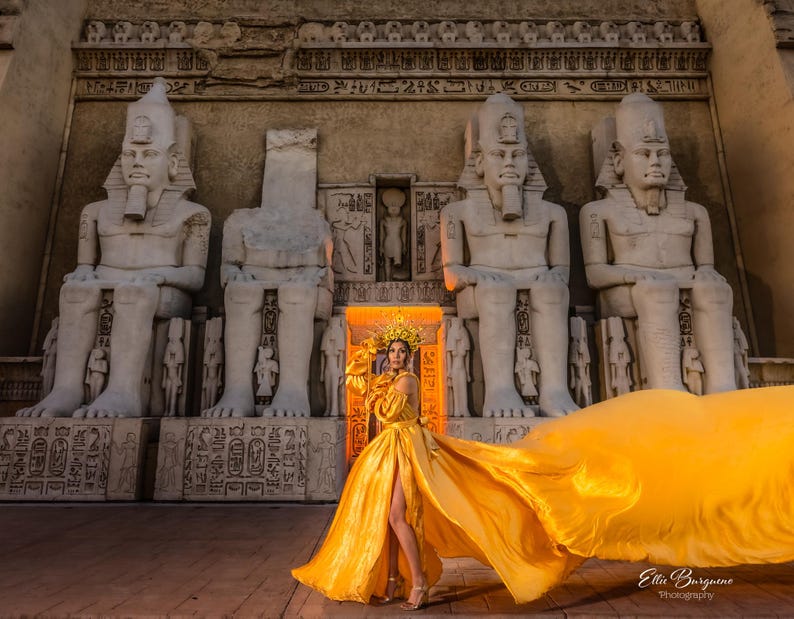 May include: A woman in a flowing gold dress and headdress stands before ancient Egyptian statues. The dress has a high slit and a long train. The background features large stone statues and hieroglyphs.