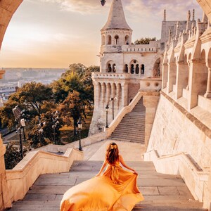 May include: A woman in a flowing, golden-yellow dress walks up a stone staircase towards a historic building with a tower. The scene is framed by an archway, with a sunset sky in the background.