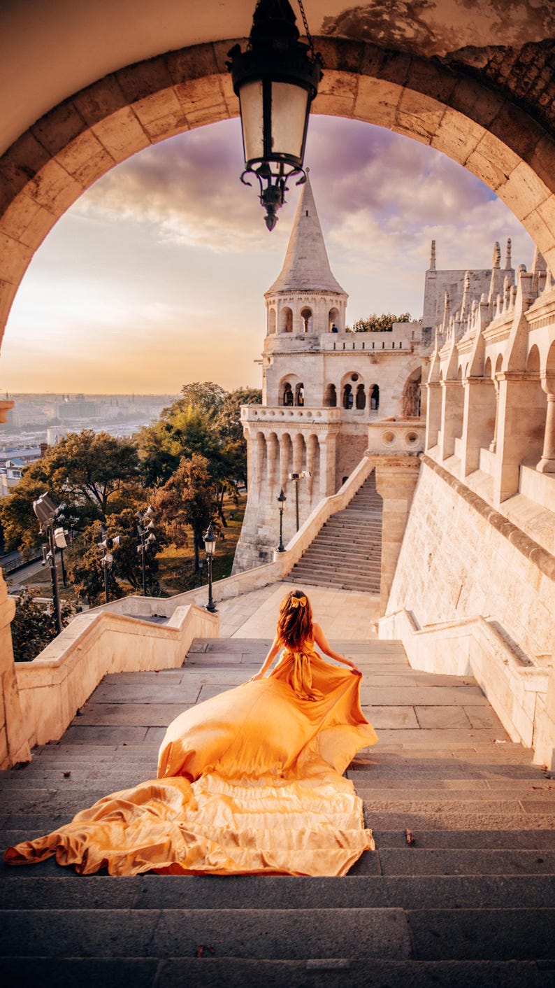 May include: A woman in a flowing, golden-yellow dress walks up a stone staircase towards a historic building with a tower. The scene is framed by an archway, with a sunset sky in the background.