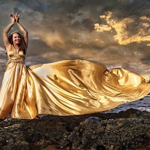 May include: A golden satin gown with a flowing train. The dress has a V-neck and a jeweled waist. The model is standing on a rocky shore with arms raised, against a dramatic sky.