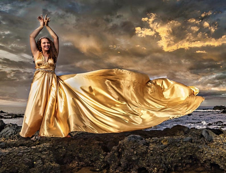 May include: A golden satin gown with a flowing train. The dress has a V-neck and a jeweled waist. The model is standing on a rocky shore with arms raised, against a dramatic sky.