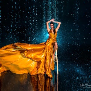May include: A woman in a flowing, golden-yellow gown poses under simulated rain. The dress features a plunging neckline and a high slit, with the fabric cascading dramatically. The background is a dark blue, creating a contrast with the dress.