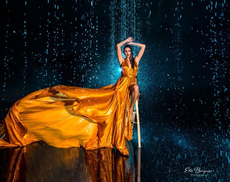 May include: A woman in a flowing, golden-yellow gown poses under simulated rain. The dress features a plunging neckline and a high slit, with the fabric cascading dramatically. The background is a dark blue, creating a contrast with the dress.