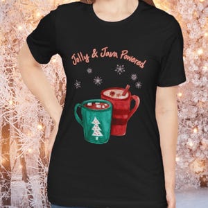 Può includere: T-shirt nero con la scritta "Jolly & Java Powered". Il design mostra due tazze di bevande calde: una tazza verde con un albero di Natale e una tazza rossa con panna.