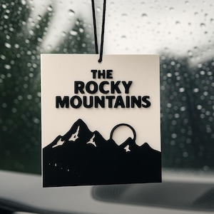 May include: A rectangular, white air freshener with the text "THE ROCKY MOUNTAINS" in bold, black letters. Below the text is a black silhouette of mountains and a sun. The air freshener hangs from a black string.