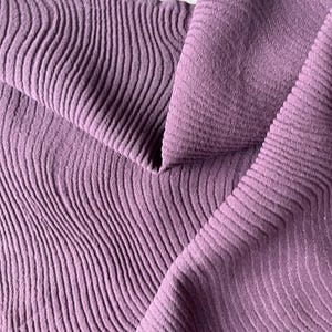 May include: Close-up of a lavender fabric with a wavy, textured pattern. The fabric is folded, revealing both the textured side and a smoother, less textured side. The background is a plain white surface.