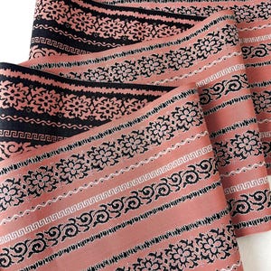 May include: Close-up of several rolls of fabric with a repeating pattern. The fabric has a salmon-colored background with black and white decorative stripes. The stripes feature floral, geometric, and scroll designs.