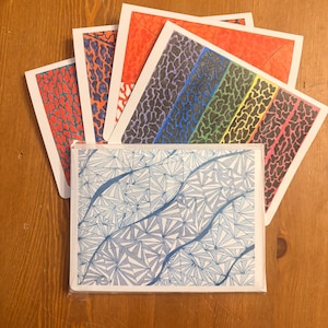 May include: A collection of greeting cards with various abstract designs. The cards feature patterns in red, orange, purple, blue, green, and pink. A stack of cards with a blue and white geometric design is in the foreground.