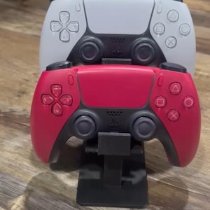 May include: A black plastic dual controller stand holding two wireless game controllers. The top controller is white with black accents, and the bottom controller is red with black accents. Both feature standard directional pads, joysticks, and buttons.