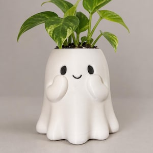 May include: A white ghost-shaped planter with a smooth finish, featuring black painted eyes and a small curved mouth. The ghost has two raised hands near its face and contains a green and yellow variegated pothos plant with several leaves.