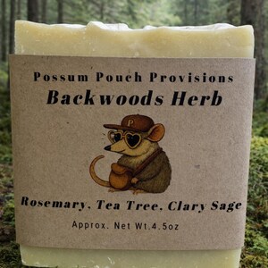 May include: A bar of Backwoods Herb soap from Possum Pouch Provisions. The soap is wrapped in a brown label featuring a cartoon possum wearing a hat and sunglasses. The label lists rosemary, tea tree, and clary sage. Net weight is approximately 4.5 oz.