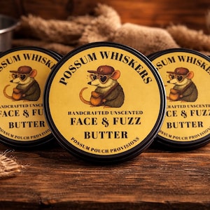 May include: Several round tins of "Possum Whiskers Face & Fuzz Butter" are arranged on a wooden surface. Each tin features a cartoon possum wearing sunglasses and a hat. The text "Handcrafted Unscented" is also visible.