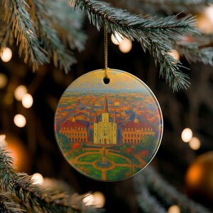 Puede incluir: Adorno navideño redondo con una pintura de una catedral y edificios circundantes. El adorno está colgado de un hilo dorado en un árbol de Navidad. La pintura presenta una catedral amarilla con una alta aguja, rodeada de edificios rojos y naranjas.