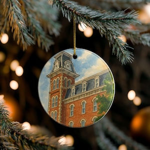 Könnte beinhalten: Runder Weihnachtsbaumschmuck aus Keramik mit einem Aquarellgemälde eines Backsteingebäudes mit einem hohen Turm und einem dunklen Dach. Der Schmuck hängt an einer goldenen Schnur vor einem verschwommenen Hintergrund aus Weihnachtslichtern und einem Tannenzweig.