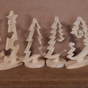 May include: Five wooden Christmas tree decorations in various designs. The trees are cut from light-coloured wood and stand on small bases. One tree has a star on top, while others have unique cutout shapes.