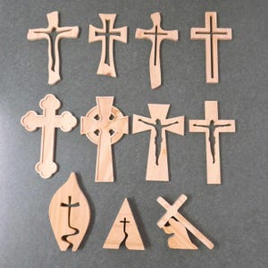 May include: A collection of eleven wooden crosses in various designs. The crosses are made of light-colored wood and feature different cut-out patterns and shapes, including a Celtic cross and a cross with a figure.
