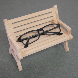 Puede incluir: Un pequeño banco de madera con asiento y respaldo de listones. Unas gafas con montura negra descansan sobre el banco. El banco es de madera clara y tiene un diseño sencillo. El fondo es una superficie gris lisa.