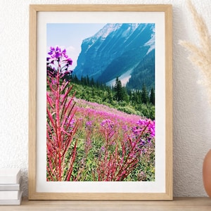 May include: A framed print featuring a vibrant mountain landscape. The foreground is filled with bright pink wildflowers, with a backdrop of a lush green hillside and a towering mountain under a blue sky. The frame is made of light-colored wood.