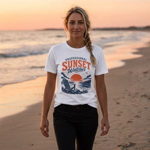 May include: White t-shirt with a graphic that reads "Professional Sunset Watcher" in orange and blue. The graphic depicts a sunset over the ocean with a beach chair. The person is wearing black pants and is standing on a beach.