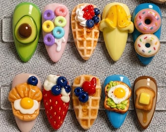 Könnte beinhalten: Zehn künstliche Fingernägel mit verschiedenen Designs zum Thema Essen. Die Designs umfassen eine Avocado, Müsli, Waffeln mit Beeren, eine Banane, Donuts, ein Croissant mit Ei, eine Erdbeere mit Schlagsahne und Blaubeeren, Waffeln mit Beeren, Toast mit Ei und Toast mit Butter.