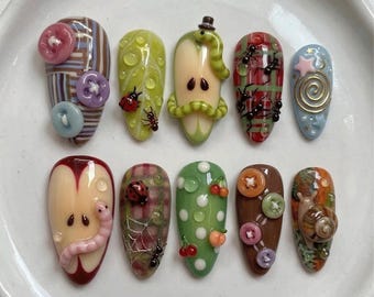 May include: A collection of ten uniquely designed nail art pieces. Each nail features a different design, including buttons, ladybugs, ants, a worm, a snail, and other whimsical elements. The color palette includes shades of brown, green, red, and blue.