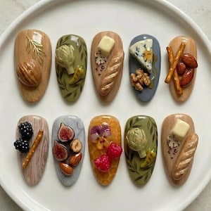 May include: A collection of ten intricately designed nail art pieces. Each nail features a different miniature food item, including nuts, artichokes, cheese, bread, berries, figs, and nuts. The nails are painted in various colors and textures, creating a unique and artistic display.