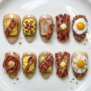 Può includere: Un set di dieci unghie finte a tema colazione. Ogni unghia presenta un diverso alimento in miniatura, tra cui toast, pancetta, uova e waffle. Le unghie sono dipinte di un colore beige neutro e sono disposte su una superficie bianca.