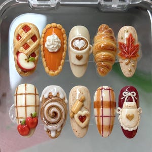 May include: A set of ten press-on nails featuring 3D autumn-themed designs including an apple pie slice, pumpkin pie with whipped cream, a coffee mug, a croissant, a maple leaf, plaid patterns, a cinnamon roll, a rolling pin, and an apron.