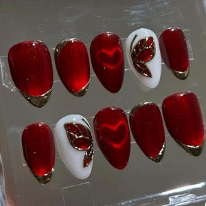 May include: Set of ten red and gold press-on nails. Some nails feature a gold trim, a heart design, or a gold butterfly design on a white base. The nails have an oval shape.