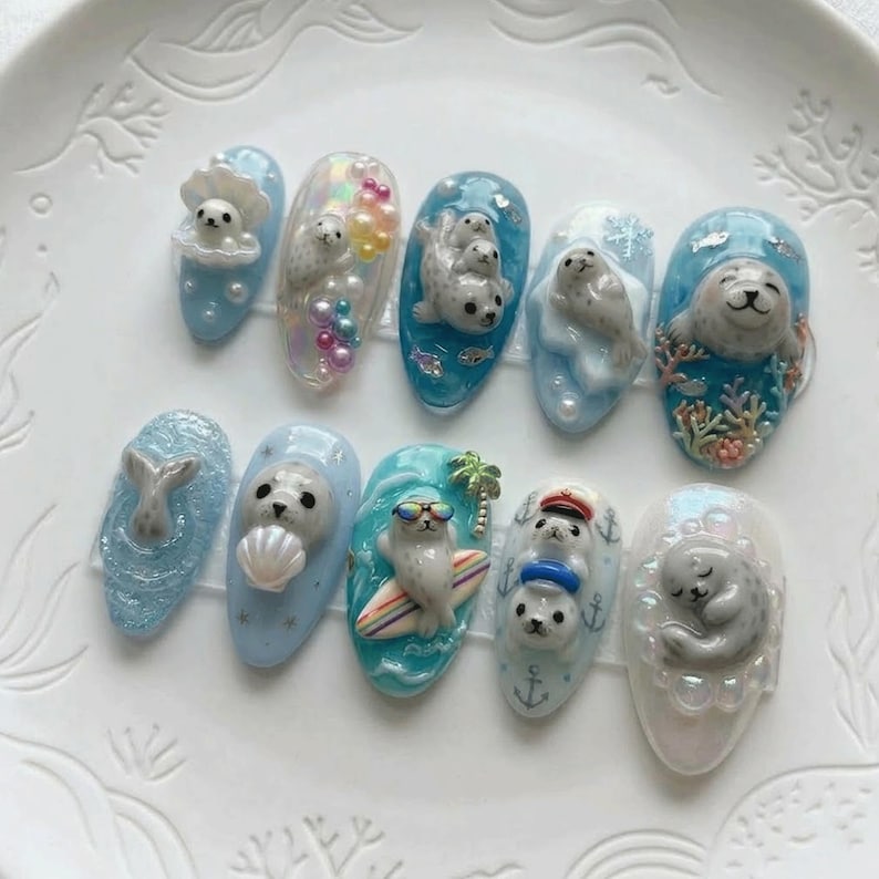 Pode incluir: Um conjunto de dez designs de nail art esculpidos com focas em v&aacute;rias cenas aqu&aacute;ticas. As unhas s&atilde;o pintadas em tons de azul e branco, com focas representadas com conchas, em pranchas de surf e em outros ambientes marinhos.