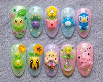 May include: A set of ten artificial nails, each with a unique design featuring colorful, cartoon-like characters and floral accents. The nails are oval-shaped and adorned with glitter and 3D embellishments, showcasing a variety of colors and textures.