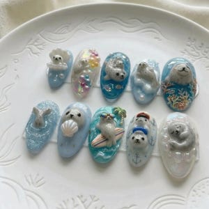 Pode incluir: Uma cole&ccedil;&atilde;o de dez designs de nail art esculpidos com focas. Cada unha apresenta uma cena diferente, incluindo focas com conchas, em pranchas de surf e em v&aacute;rios ambientes aqu&aacute;ticos. As unhas s&atilde;o pintadas em tons de azul, branco e cores iridescentes.