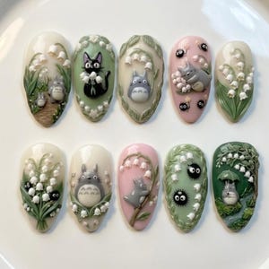 Puede incluir: Un conjunto de diez dise&ntilde;os de u&ntilde;as en forma de almendra. Cada u&ntilde;a presenta un dise&ntilde;o diferente, incluyendo los personajes de la pel&iacute;cula de animaci&oacute;n "Mi Vecino Totoro", con flores de lirio de los valles y escenas de bosque. La paleta de colores incluye blanco, verde, rosa y gris.