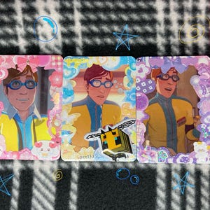 Könnte beinhalten: Drei Sammelkarten mit einer Zeichentrickfigur in einem gelb-blauen Outfit, die eine blaue Brille trägt. Jede Karte ist mit einem dekorativen Rahmen in Rosa, Blau und Lila versehen, mit zusätzlichen Verzierungen. Eine Karte hat eine pixelige Bienen-Grafik.