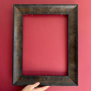 Pode incluir: Uma moldura retangular marrom escuro com um acabamento desgastado. A moldura é exibida em um fundo vermelho. A moldura tem uma borda costurada e uma aparência ligeiramente desgastada, sugerindo um estilo antigo ou vintage.