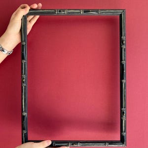 Puede incluir: Un marco de fotos rectangular negro con un diseño de estilo bambú se sostiene sobre un fondo rojo. El marco tiene un acabado oscuro con sutiles detalles dorados y está sostenido por dos manos.