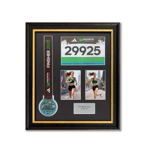 Op de afbeelding: Ingelijste Manchester Marathon 2023 herdenkingsstukken. Inclusief een medaille, een startnummer met nummer 29925, twee foto's van een hardloper en een plaatje met de finishtijd. De lijst heeft een zwarte rand met een gouden binnenrand.