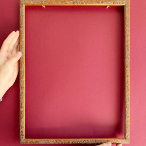 May include: A rectangular, ornate, gold-colored picture frame with detailed carvings. The frame is held by two hands against a solid red background. The frame is empty, ready for a photo or artwork.