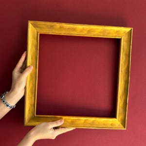 May include: A gold-colored, square picture frame with a textured surface. The frame is held against a deep red background. The frame is empty, ready for a photo or artwork.