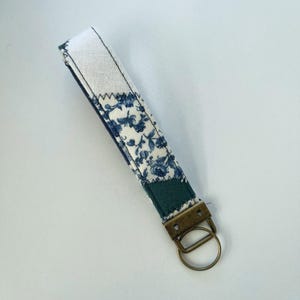 May include: A fabric wristlet keychain featuring a white and blue floral pattern on one side and a solid dark teal fabric on the other. The strap is secured with a bronze-toned metal hardware clamp and a circular split ring for attaching keys.