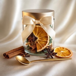 May include: A glass jar filled with dried orange slices, star anise, and cinnamon sticks, tied with a cream-colored ribbon. A gold spoon and additional spices are arranged nearby, creating a warm, inviting aesthetic.